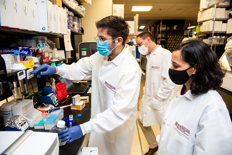 Three scientists wearing lab coats are standing near a lab bench and looking at a tool. 