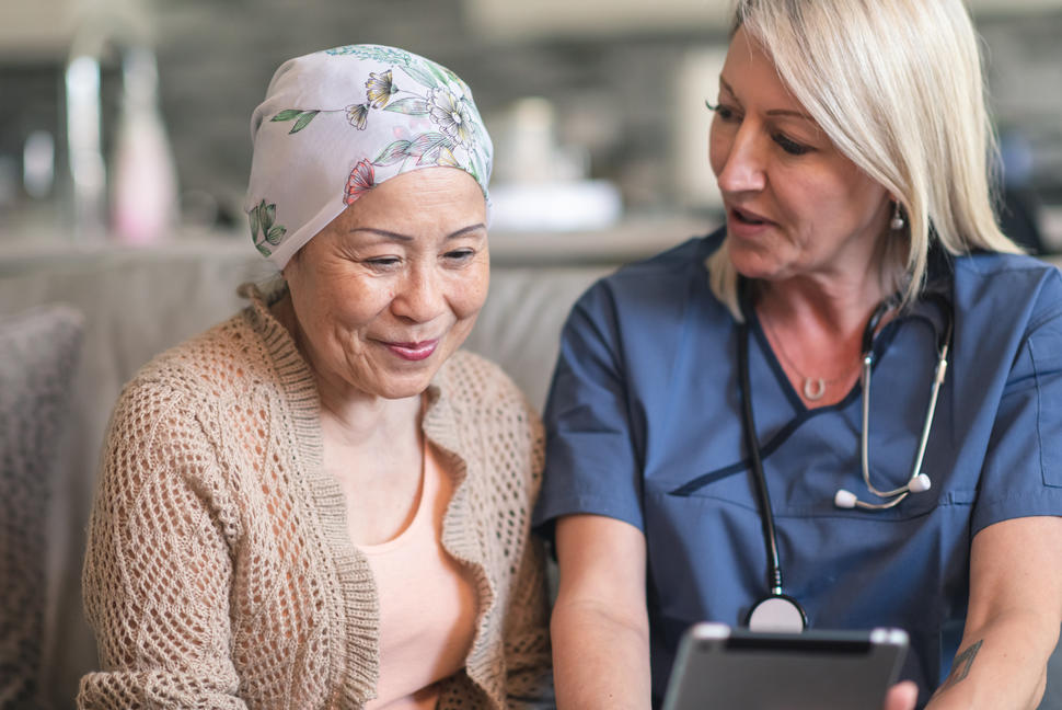 A female patient wearing a headscarf sits next to a female physician. The physician is showing the patient a wireless tablet.