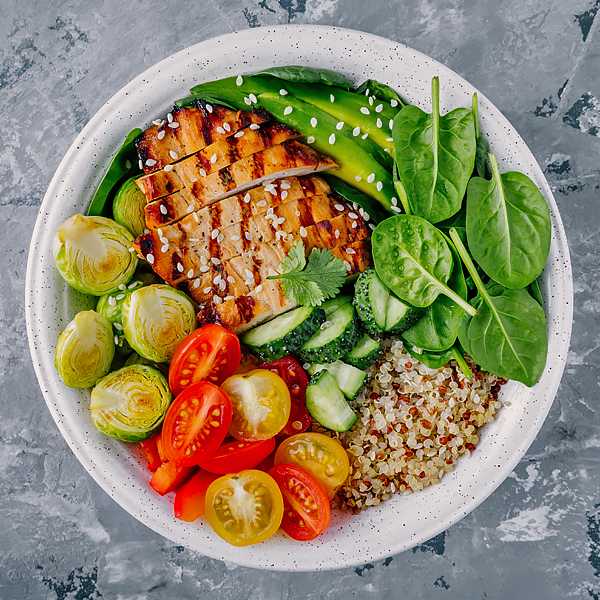 A healthy bowl of food with grilled chicken, avocado, spinach, quinoa, cucumbers, tomatoes, and brussels sprouts on a grey background