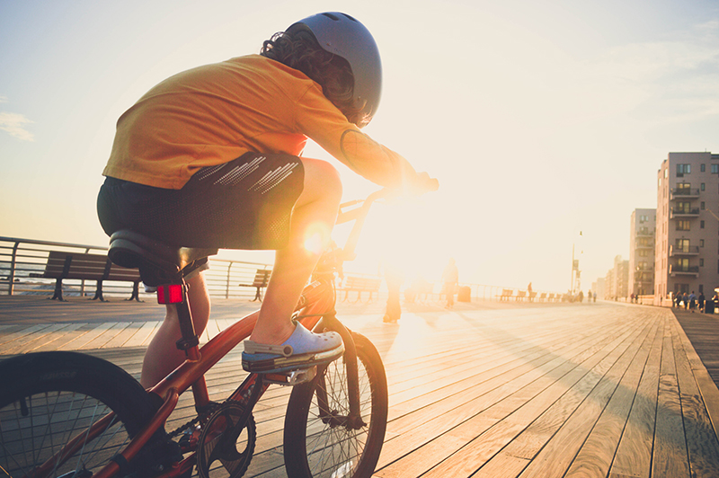 A young boy riding his bicycle on the boardwalk by the beach backlit by the sun wearing a helmet.