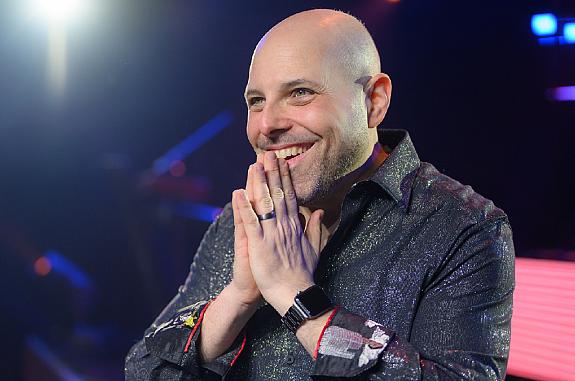 Cancer survivor stands on a stage with hands clasped together and a smile on his face.