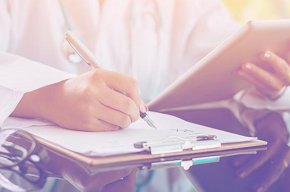 Person wearing a lab coat sitting at a table holding an electronic tablet and writing on a clipboard.