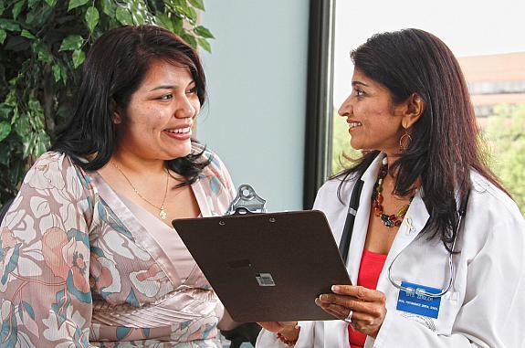 A female doctor holding a clipboard talks with a Hispanic female patient.