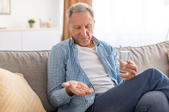 Older man sitting on a couch with a glass of water, looking at pills in his hand
