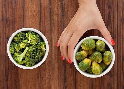 A woman’s hand holding a bowl of brussel sprouts, with a bowl of cooked broccoli to the left. A woman’s hand holding a bowl of brussel sprouts, with a bowl of cooked broccoli to the left.