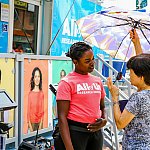 Young woman with All of Us tee shirt speaks to woman holding an umbrella.