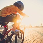 A young boy riding his bicycle on the boardwalk by the beach backlit by the sun wearing a helmet.