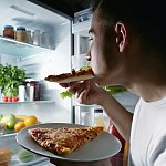 Young man eating pizza from a refrigerator at night.